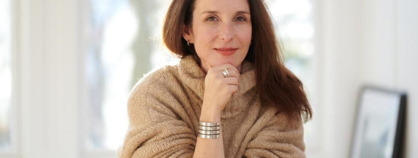 A woman with long brown hair hanging over her left shoulder sits on a chair with her right arm under her chin. She wears a brown sweater and sits in a bright white room.