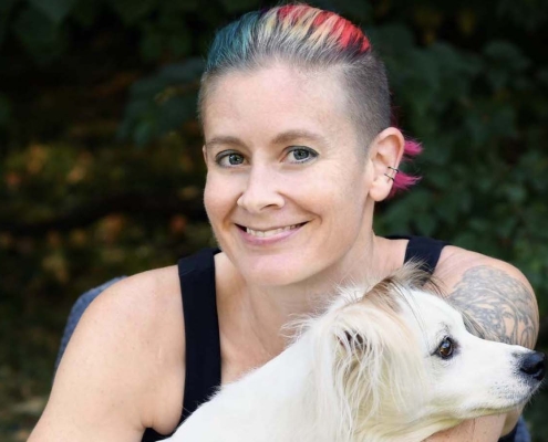 A person with rainbow colored hair that are shaved on the sides. The person wears a black tank top and holds a fluffy white dog.