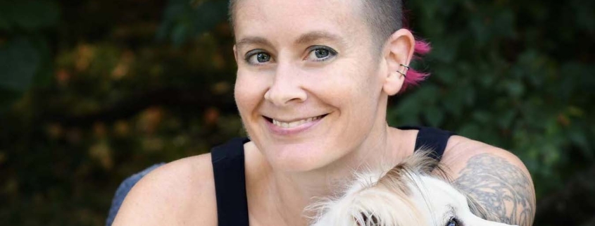 A person with rainbow colored hair that are shaved on the sides. The person wears a black tank top and holds a fluffy white dog.