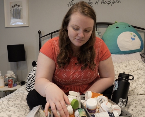 Sarah, a woman with long blonde hair sits on her bed with a big box of medication in front of her.