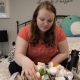 Sarah, a woman with long blonde hair sits on her bed with a big box of medication in front of her.