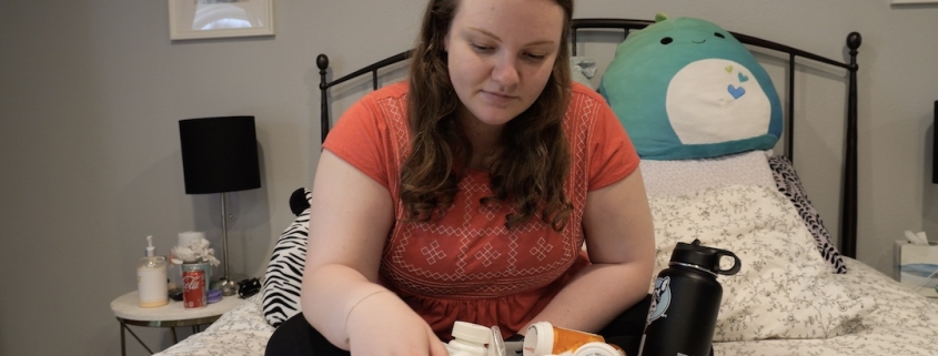 Sarah, a woman with long blonde hair sits on her bed with a big box of medication in front of her.