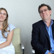 Two people are sitting in beige office chairs in front of a white wall. Dr. Gensemer is a woman with long, wavy blonde hair. She wears a blue shirt and white pants and has flower tattoos on her arm. Next to her sits Dr. Norris, a man with short brown hair, grey glasses, a blue shirt and a dark blue jacket on top.