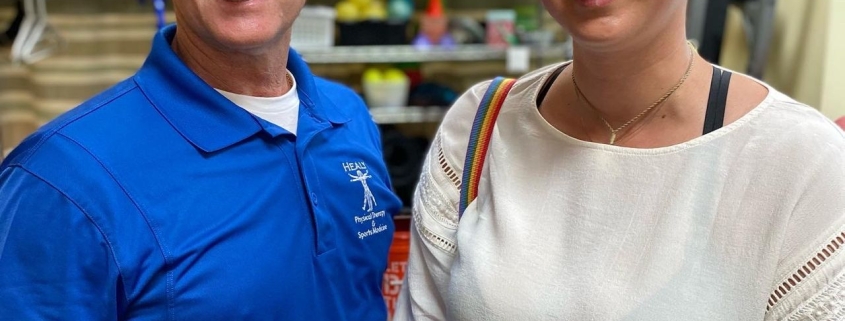 Two people in a physical therapy office standing next to each other. A woman with short brown hair and a white shirt and pink skirt smiles brightly. Next to her stands a man with a blue shirt and a stethoscope around his neck who also smiles brightly.