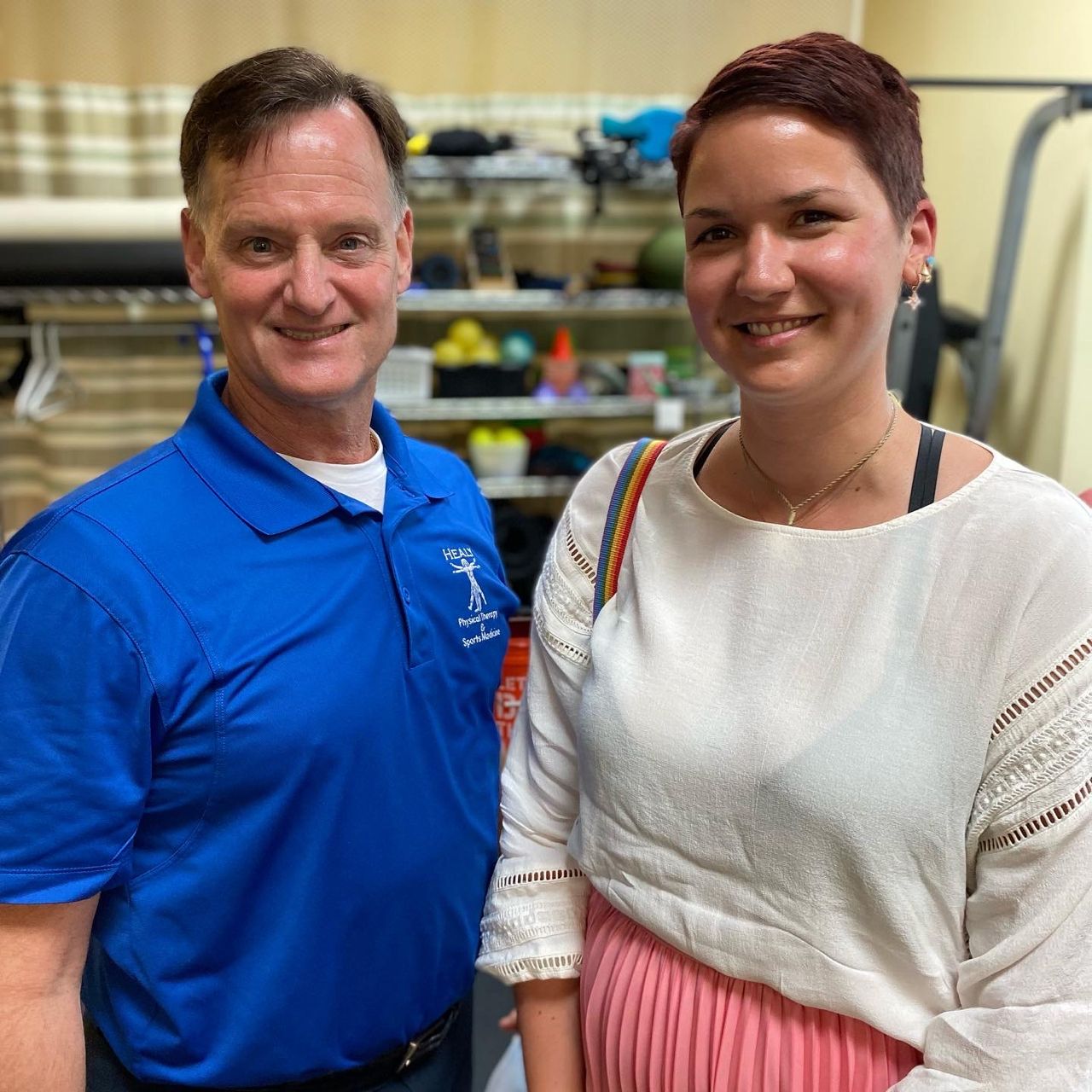 Two people in a physical therapy office standing next to each other. A woman with short brown hair and a white shirt and pink skirt smiles brightly. Next to her stands a man with a blue shirt and a stethoscope around his neck who also smiles brightly.