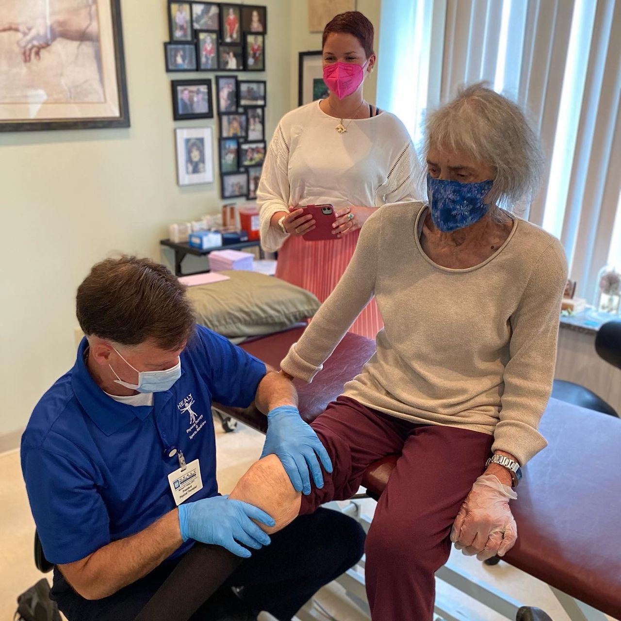 Three people in a physical therapy office. A woman with grey hair and a blue face mask sits on a treatment bench while a man with a blue shirt and a stethoscope around his neck treats her knee. In the background is a woman with short brown hair and a white shirt and pink skirt taking photos.