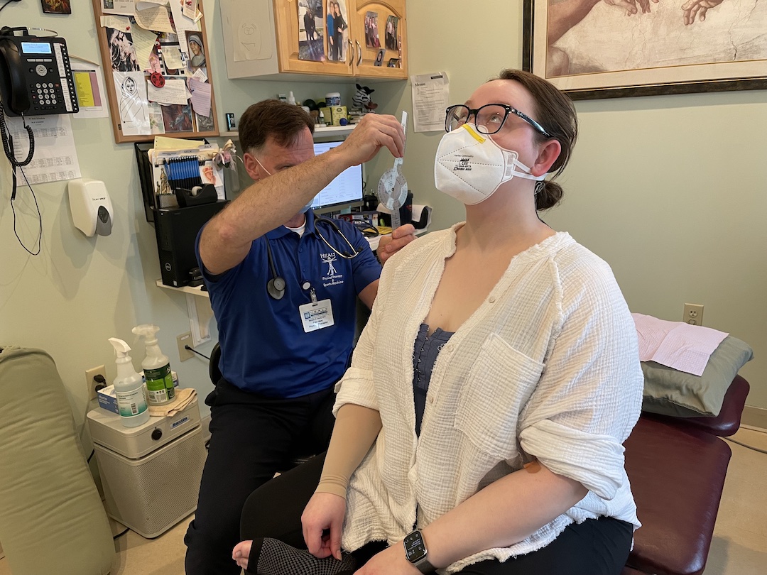 Two people in a physical therapy office. A woman sits on a treatment bench, wearing a white face mask, a white shirt and black glasses. She extends her head backwards and looks to the ceiling. The man next to her wears a blue shirt and a stethoscope around his neck and measures her neck movement.