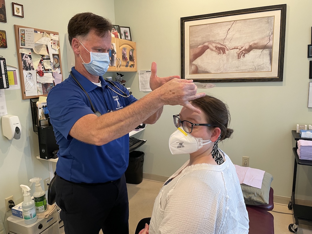 Two people in a physical therapy office. A woman sits on a treatment bench, wearing a white face mask, a white shirt and black glasses. She rotates her head to the left . The man standing in front of her wears a blue shirt and a stethoscope around his neck and has has hands on her forehead.