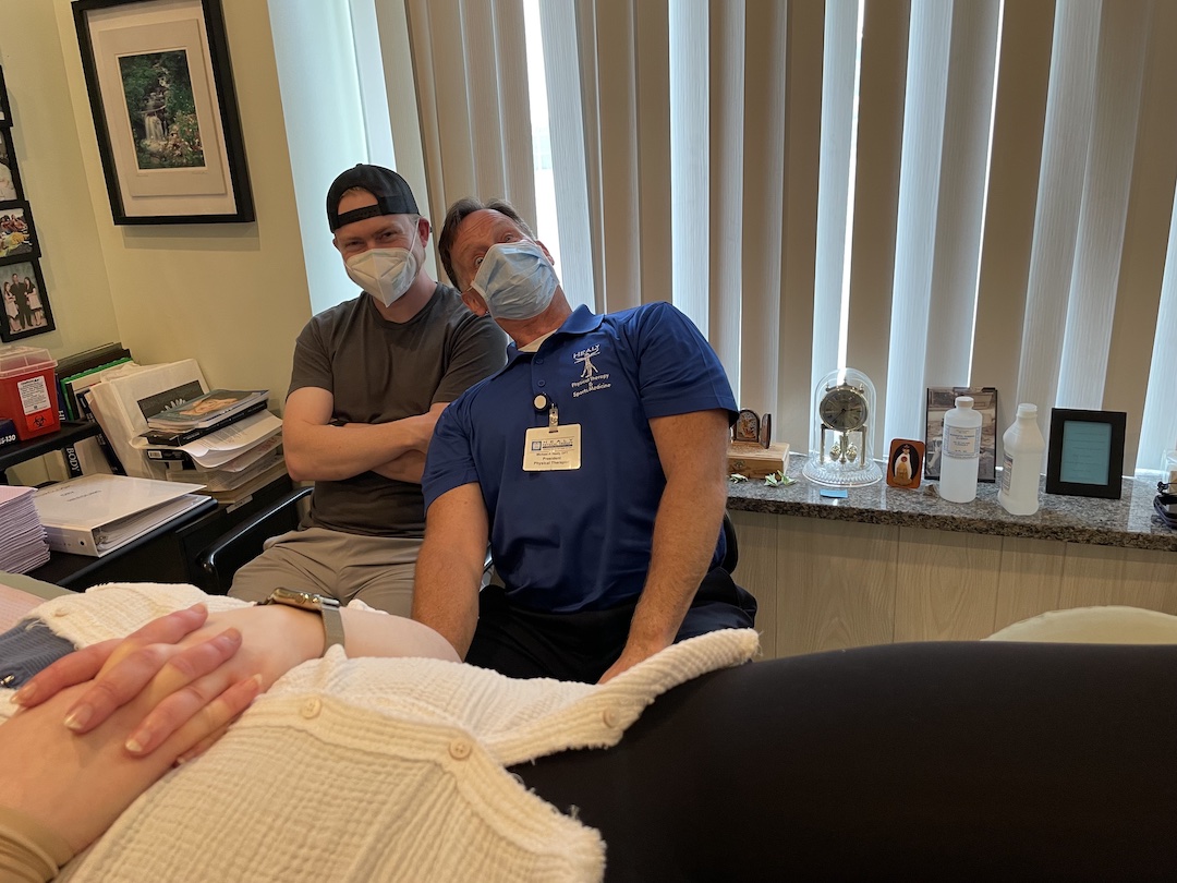 Three people in a physical therapy office. A woman lies on a treatment bench, wearing a white face mask, a white shirt and black glasses. Two men are next to her goofing around. One wears a blue shirt and a stethoscope around his neck and leans back next to another man with a black hat and a white face mask. They smile under their masks.