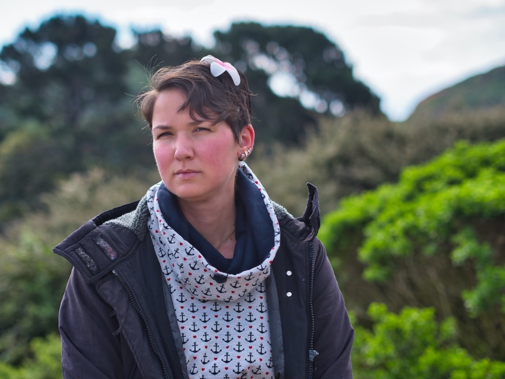 Karina, eine Frau mit kurzen, braunen Haaren und einer Blume im Haar sitzt in der Natur. Im Hintergrund sieht man unterschiedlich grüne Büsche und Bäume.