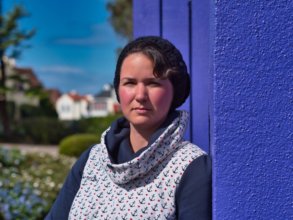 Karina, eine Frau mit kurzen braunen Haaren steht an einer lilafarbenen Wand und blickt in die Ferne. Im Hintergrund sieht man bunte Blumen, Häuser und einen strahlend blauen Himmel.