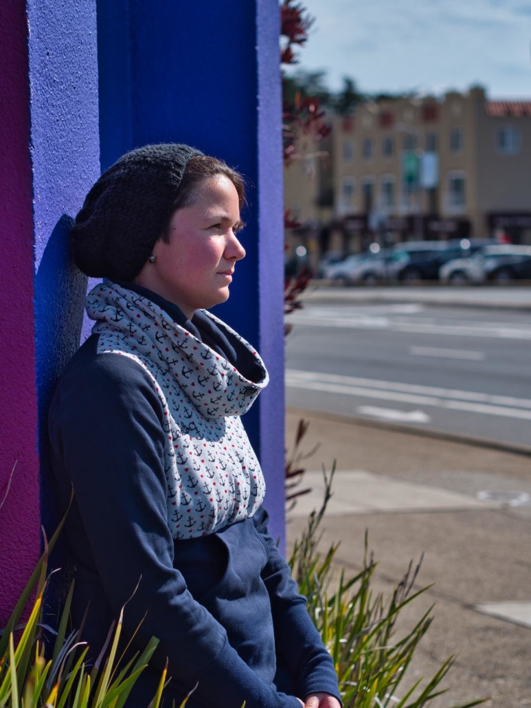 Karina, eine Frau mit kurzen braunen Haaren steht an einer lilafarbenen Wand und blickt in die Ferne. Im Hintergrund sieht man eine Straße und parkende Autos.