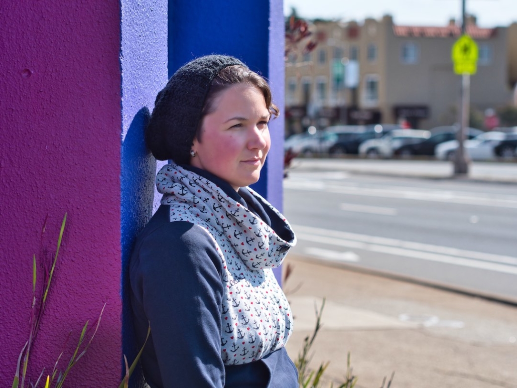 Karina, eine Frau mit kurzen braunen Haaren steht an einer lilafarbenen Wand und blickt in die Ferne. Im Hintergrund sieht man eine Straße und parkende Autos.