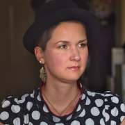 A woman with short brown hair and a black hat sits in a kitchen. She wears wing-like earrings and a polkadot shirt.