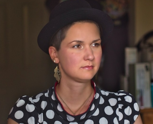 A woman with short brown hair and a black hat sits in a kitchen. She wears wing-like earrings and a polkadot shirt.