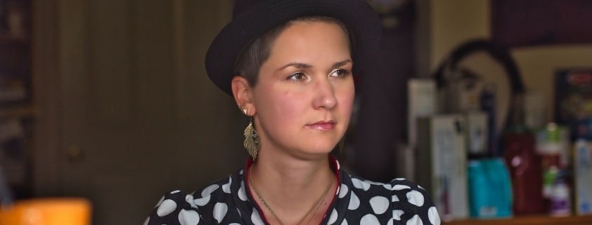 A woman with short brown hair and a black hat sits in a kitchen. She wears wing-like earrings and a polkadot shirt.