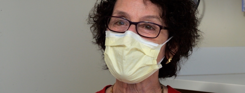 Dr. Wolf, a woman with short, curly brown hair, black glasses and a yellow face mask.