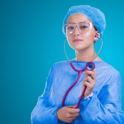 A female doctor with a blue surgical robe and a blue cap holding up a purple stethoscope.
