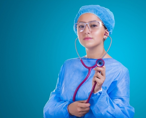 A female doctor with a blue surgical robe and a blue cap holding up a purple stethoscope.