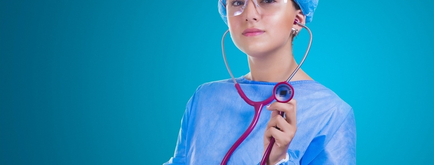 A female doctor with a blue surgical robe and a blue cap holding up a purple stethoscope.