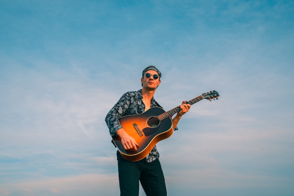 Marvin, a man with a hippie outfit and an acoustic guitar stands outdoors with a bright blue sky in the background.