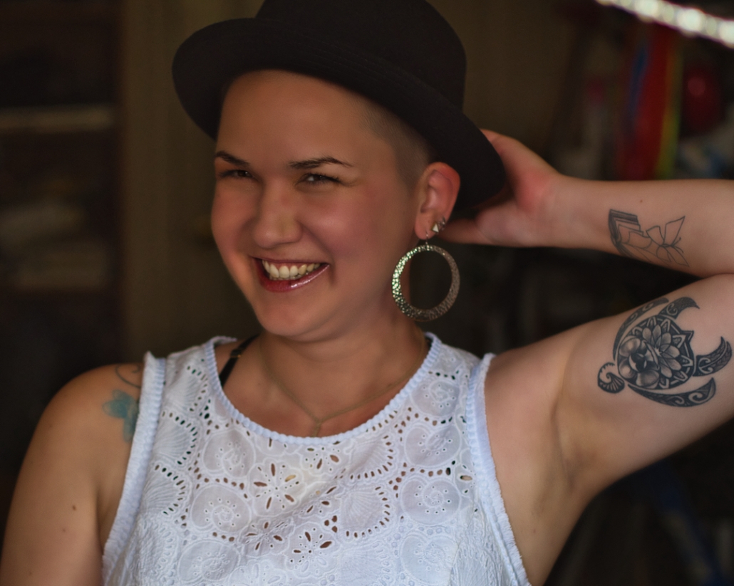 Karina, a woman with a shaved head wears a white delicate dress and a black hat with huge silver earrings.