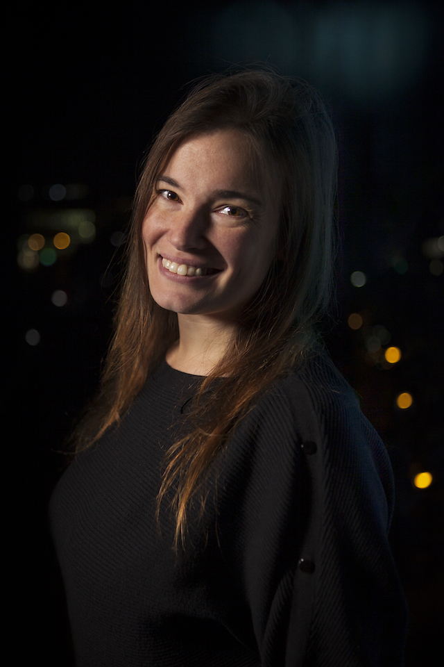 Dhana, a woman with long brown hair and a black shirt stands outside in front of a dark city landscape.