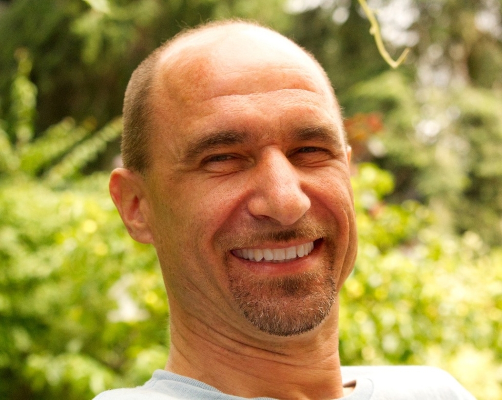 Stefan, a bald man with a beard smiles brightly with many green bushes in a blurred background