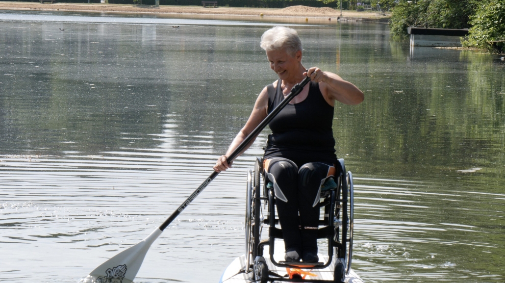 Rita, eine Frau mit kurzen grauen Haaren, sitzt in einem Rollstuhl, der an einem Stand Up Paddle Board auf einem See befestigt ist. Es ist sonnig und hell und sie lächelt, während sie ein Paddel hält.