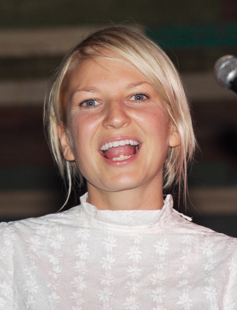 Sia, a woman with blonde hair and a white shirt with her mouth open as if she was singing.