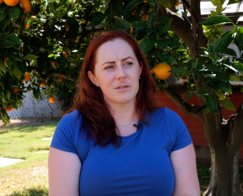 Caitlin, a woman with long red hair is sitting on a chair in her backyard. The sun is shining. She wears a blue shirt and sits in front of a gigantic orange tree with lots of oranges and green leaves.