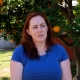 Caitlin, a woman with long red hair is sitting on a chair in her backyard. The sun is shining. She wears a blue shirt and sits in front of a gigantic orange tree with lots of oranges and green leaves.
