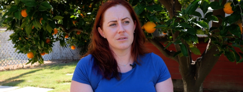 Caitlin, a woman with long red hair is sitting on a chair in her backyard. The sun is shining. She wears a blue shirt and sits in front of a gigantic orange tree with lots of oranges and green leaves.