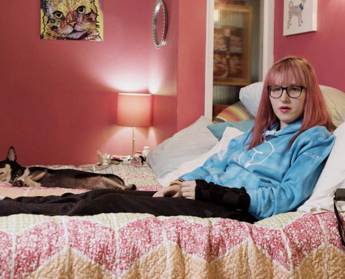 The picture shows a young woman with straight, shoulder-length pink hair and glasses, sitting on a bed. She is wearing a light blue hoodie and black pants. To her right, there is a dog lying on the bed. The room has a red wall behind the bed, and there is a framed picture of a cat above the bed. On the left side of the bed, there is a small round mirror on the wall and a bedside table with a lamp. The bedspread is pink and white with a floral pattern.