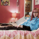 The picture shows a young woman with straight, shoulder-length pink hair and glasses, sitting on a bed. She is wearing a light blue hoodie and black pants. To her right, there is a dog lying on the bed. The room has a red wall behind the bed, and there is a framed picture of a cat above the bed. On the left side of the bed, there is a small round mirror on the wall and a bedside table with a lamp. The bedspread is pink and white with a floral pattern.