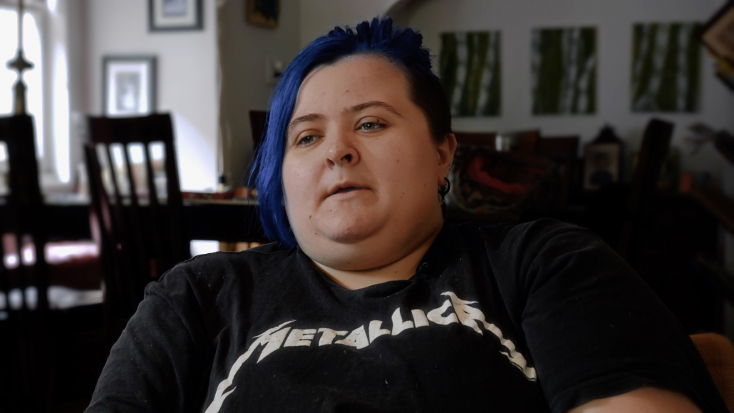 Jack, eine junge Person mit blauem, zu einem Pferdeschwanz gebundenem Haar, sitzt in einem Wohnzimmer. Sie tragen ein Metallica-T-Shirt.