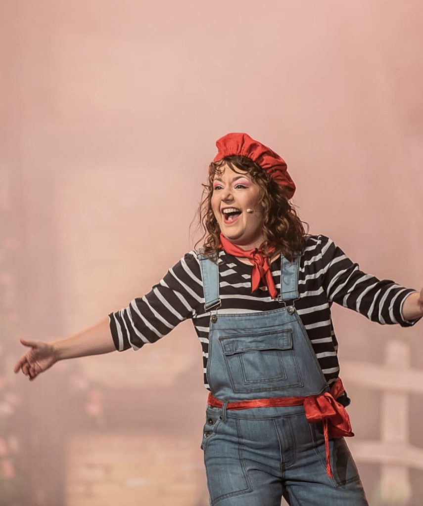 Katie Pritchard performing as Crepe Suzette in Beauty and the Beast at Worthing Pavilion Theatre. She is wearing a red beret with a red neckerchief and belt. Along with denim dungarees and a navy and white striped t-shirt.