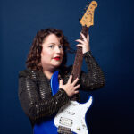 A photo of me in a gold sequinned dress clutching my Yamaha Pacifica Electric Guitar very closely