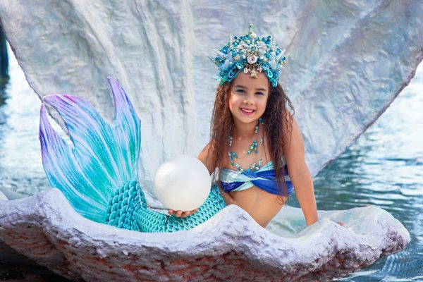 A beautiful little girl in a mermaid costume sits in a huge shell and holds a large pearl