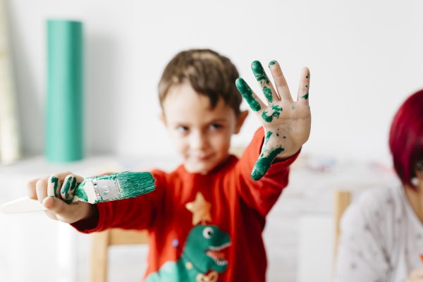 Boy showing a brush in one hand and the other painted green while doing crafts at home