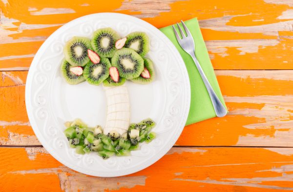 Fruit dessert for kids with kiwi, banana and strawberry on wooden background. Palm tree from fruit