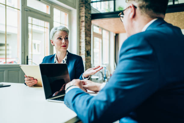 Shot of a two confident business persons sitting on a conference table in the office. Businessman and businesswoman in meeting discussing business strategy, using laptop and digital tablet. Business coworkers working together in the office.