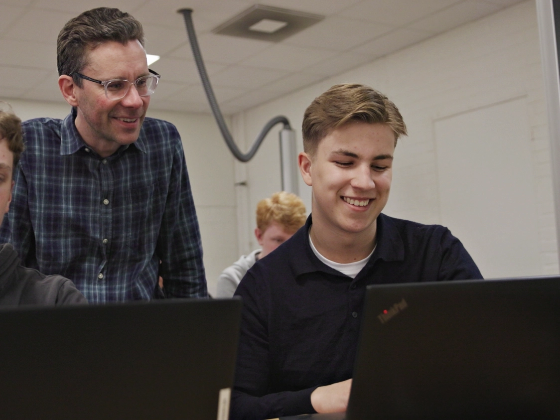 Lärare som engagerar sig med en student som arbetar vid en laptop och spelar klimatspelet i ett klassrum, båda ler.