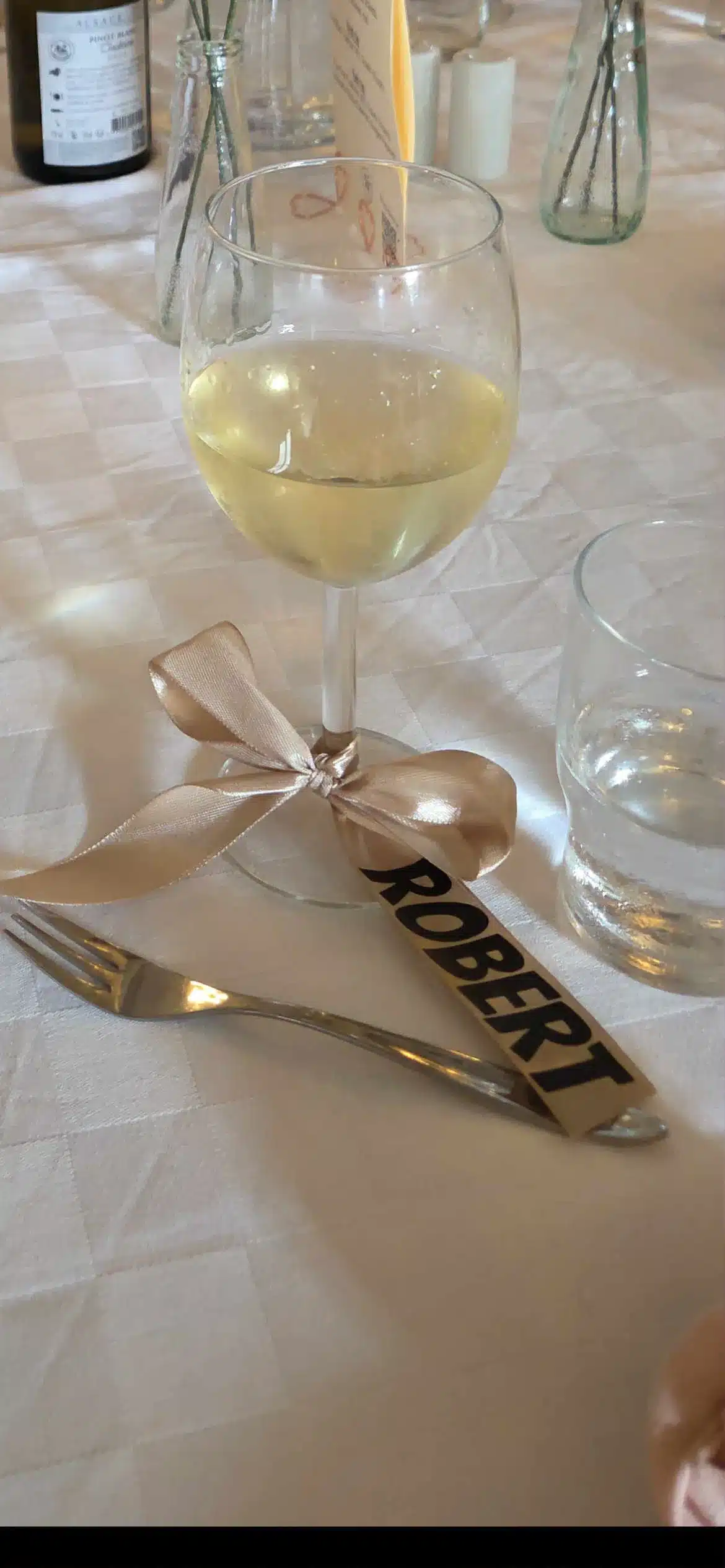 Nærbillede af et hvidvinsglas på en hvid dug, hvor et champagnefarvet silkebånd med navnet Robert er bundet om stilken som et personligt bordkort lavet med sort strygevinyl.