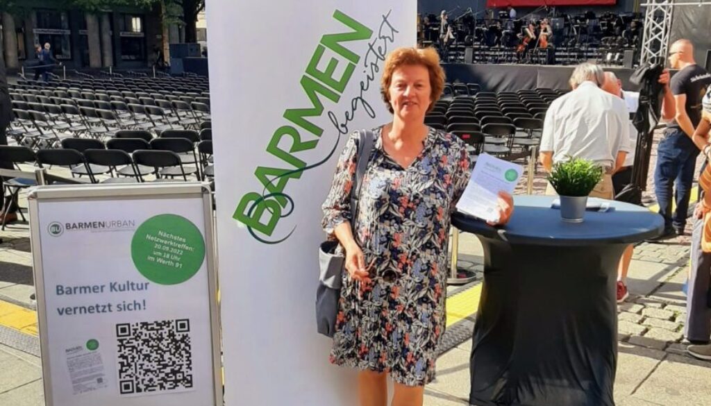 2022 Dörte Bald beim Spielzeitauftakt Wuppertaler Bühnen 2022 Dörte Baldsteht neben Aufsteller mit Netzwerkinformationen, 'Barmen besteister' Plakat und Stehtisch, im Hintergrund eine Bühne mit Stühlen auf dem Johannes-Rau-Platz