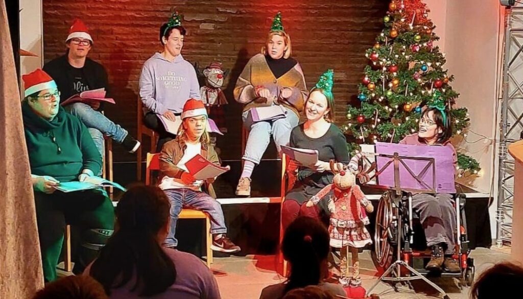 Kulturadvent 2022 Integratives Schauspielstudio im KuKi Cafe Eine Gruppe von Menschen sitzt mit Weihnachtsmützen neben einem geschmückten Tannenbaum, im Publikum sitzen Kinder