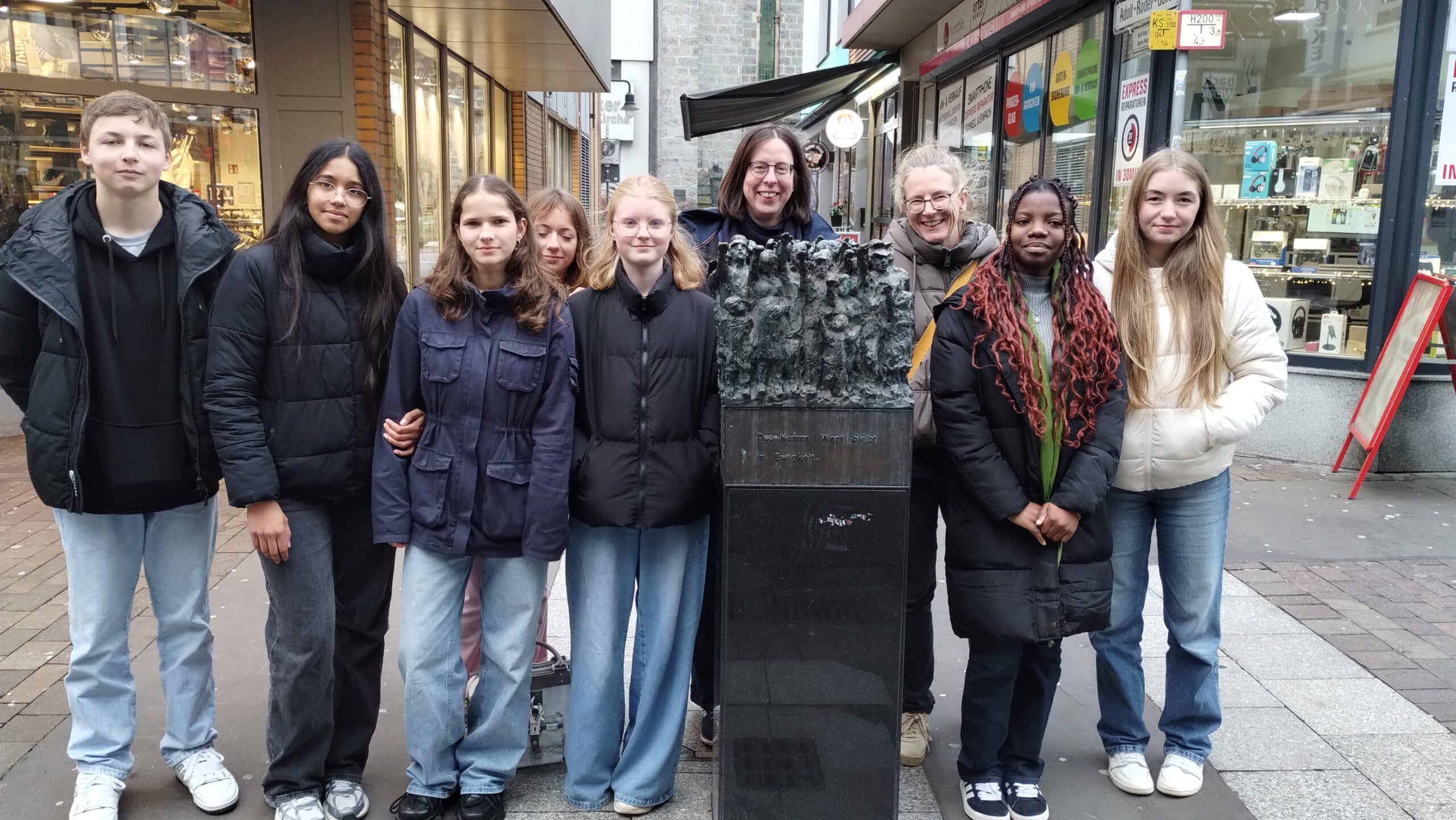 Kulturadvent 2024-12-10 Ankündigung Denkmal nach @Frank Grünberg, Kirchliche Hochschule Wuppertal Acht Jugendliche und eine Frau stehen vor dem Ulle-Hees Denkmal in Wuppertal Barmen