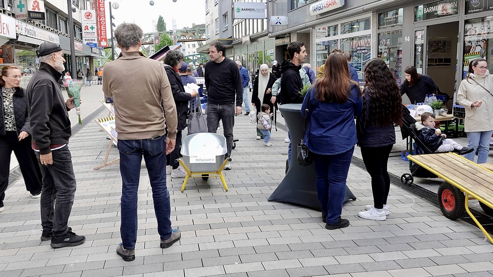 Menschen in einer Fußgängerzone, Liegestähle, eine Schubkarre, Getränketisch. Im Hintergrund das Gerüst der Wuppertaler Schwebebahn