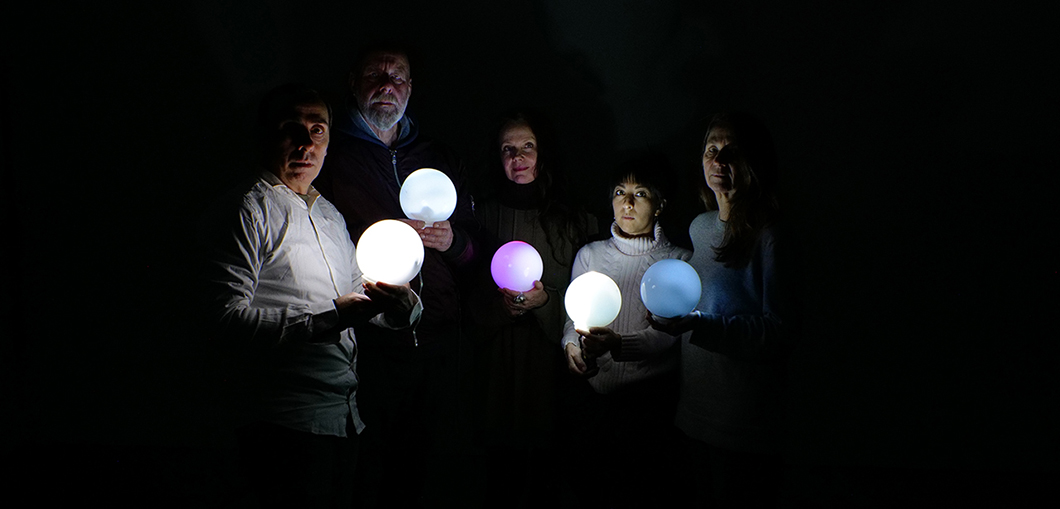 Five people are standing in the middle of a completely dark room. Each person is holding a glowing, round lamp in front of them, so only their faces and upper bodies are visible.