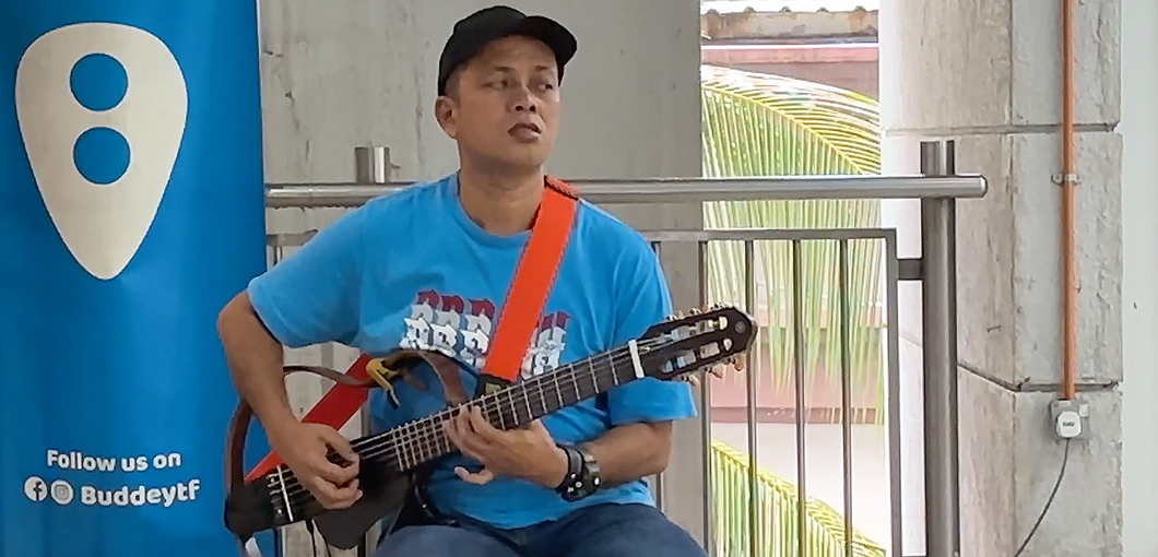 A blind man is sitting on a chair at a train station, playing guitar. The man is wearing a t-shirt, jeans, and a cap. The body of the guitar consists only of an outer frame, as it is electric and does not need a physical body to play. This is a still image from the short film 'Invisible Music.'